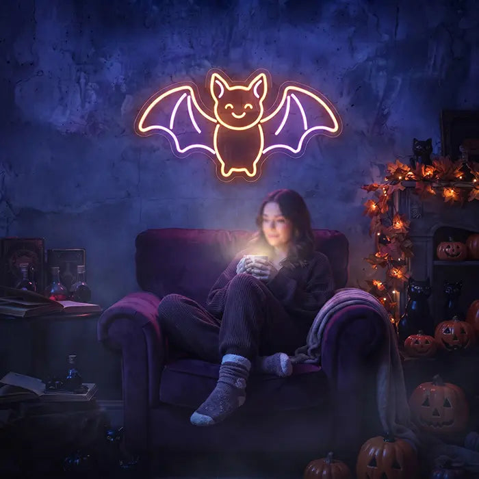 Fledermaus Neon LED Schild leuchtet über einem Sessel mit Frau und Kürbissen bei Halloween-Deko