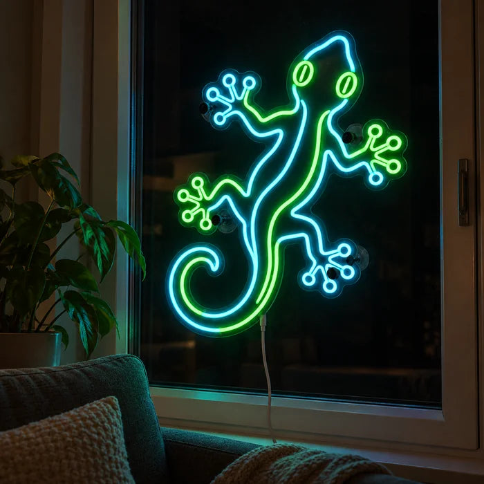 Grüner Gecko Neon LED Schild abends im Fenster eines gemütlichen Wohnzimmers