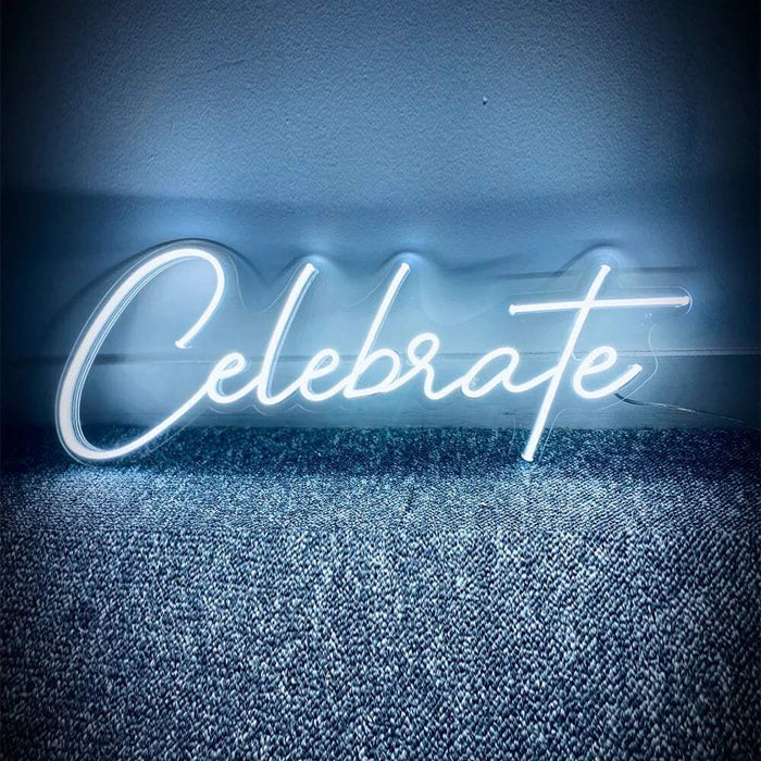 Celebrate Neon LED Schild in weißer Schreibschrift, leuchtend an eine Wand gelehnt auf grauem Teppichboden