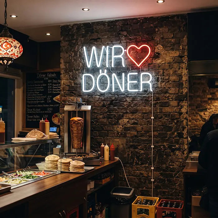 Wir lieben Döner Neon LED Schild an einer Backsteinwand im Imbiss, darunter Theke mit Dönerspieß