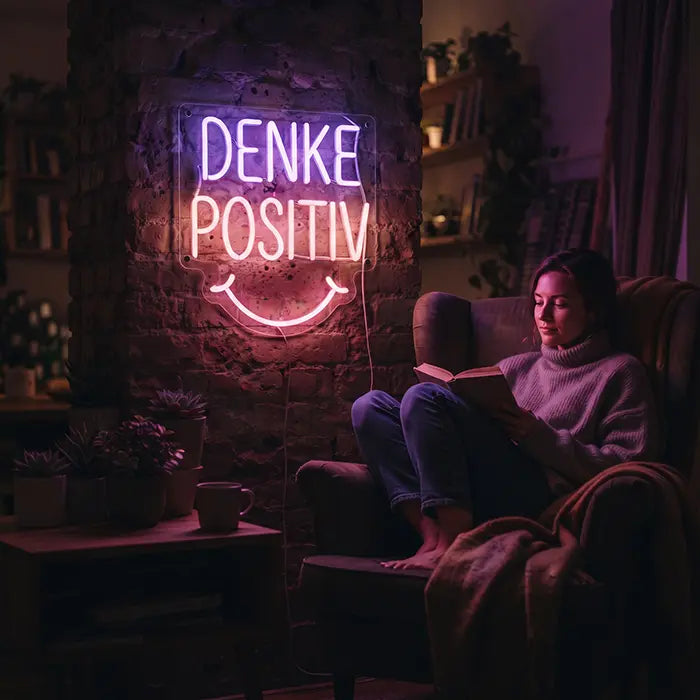 Denke Positiv Neon LED Schild leuchtet in einer Leseecke, in der eine Frau im Sessel ein Buch liest