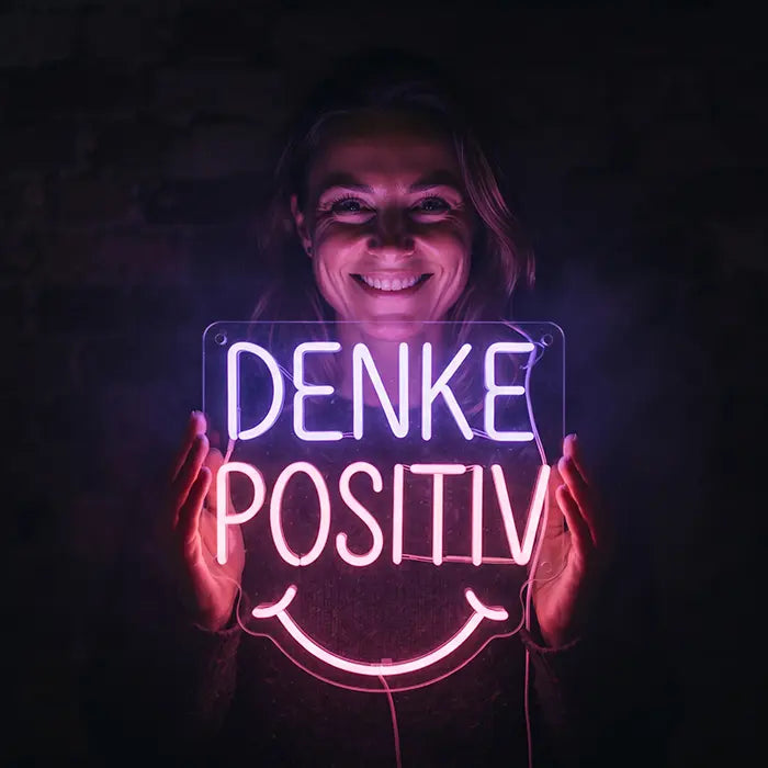 Denke Positiv Neon LED Schild wird von einer lächelnden Frau vor einer dunklen Backsteinwand gehalten