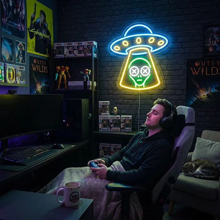 UFO Alien Entführung Neon LED Schild über einem Gaming-Setup mit Sammelfiguren, davor eine Person mit Headset