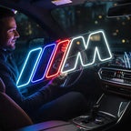 Leuchtendes Neon-Schild mit dem BMW M Logo in den charakteristischen Farben Blau, Violett und Rot sowie einem weißen "M", platziert auf einem Holzschrank